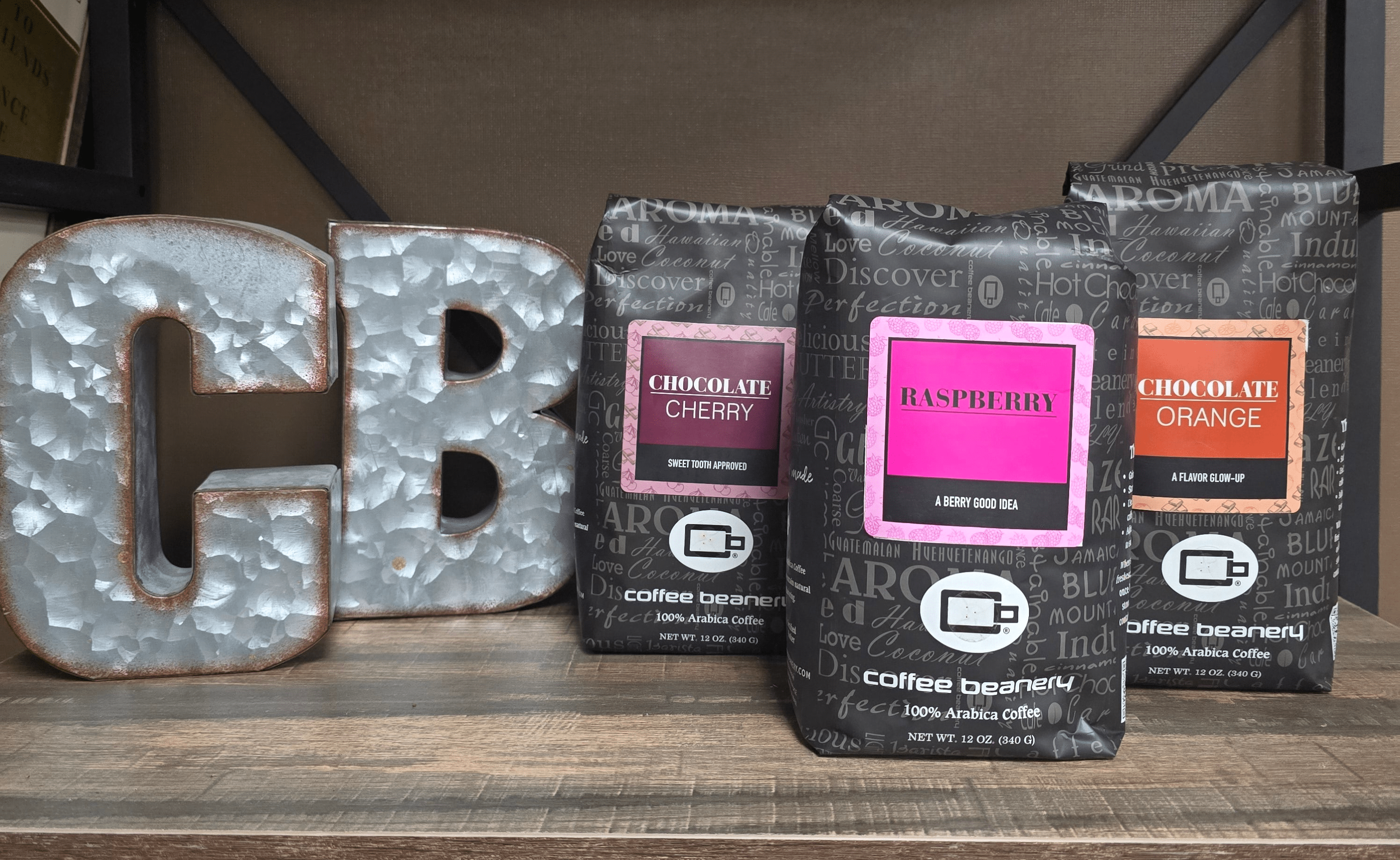 Three coffee packages with flavors 'Chocolate Cherry', 'Raspberry', and 'Chocolate Orange' on a wooden surface with decorative metal letters.